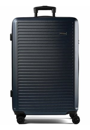 Grande valise rigide bleu foncé avec des rainures horizontales, poignée rétractable et quatre roues noires pivotantes.