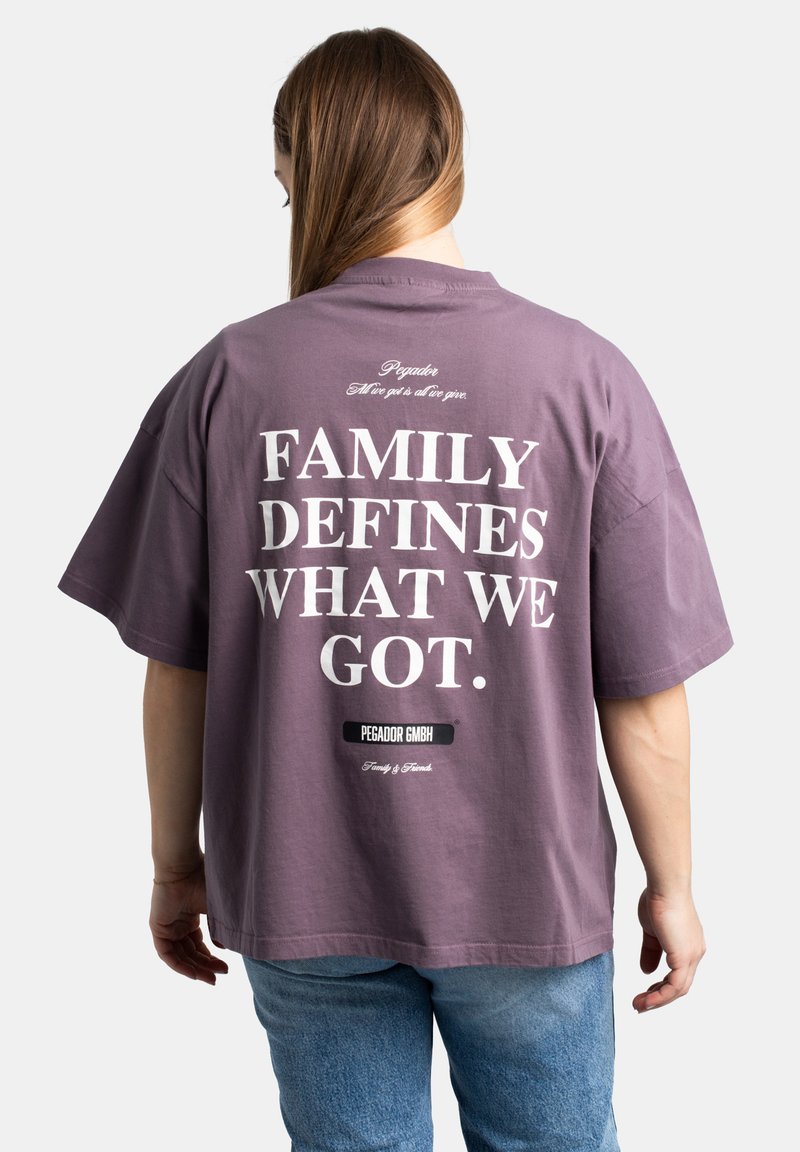 Camiseta de corte holgado en algodón morado apagado. La parte posterior presenta un gran texto blanco: "LA FAMILIA DEFINE LO QUE TENEMOS." con la marca debajo. Textura suave, mangas cortas.