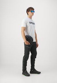 Czarny kask trzymany w ręku, biały t-shirt z czarnym logo, czarne spodnie z zielonymi suwakami i czarne buty z zielonymi akcentami.
