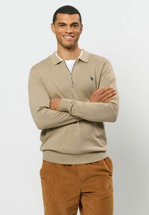 Homme aux cheveux courts et foncés, souriant, les bras croisés, portant un pull beige à col zippé et un pantalon en velours côtelé marron sur un fond uni.