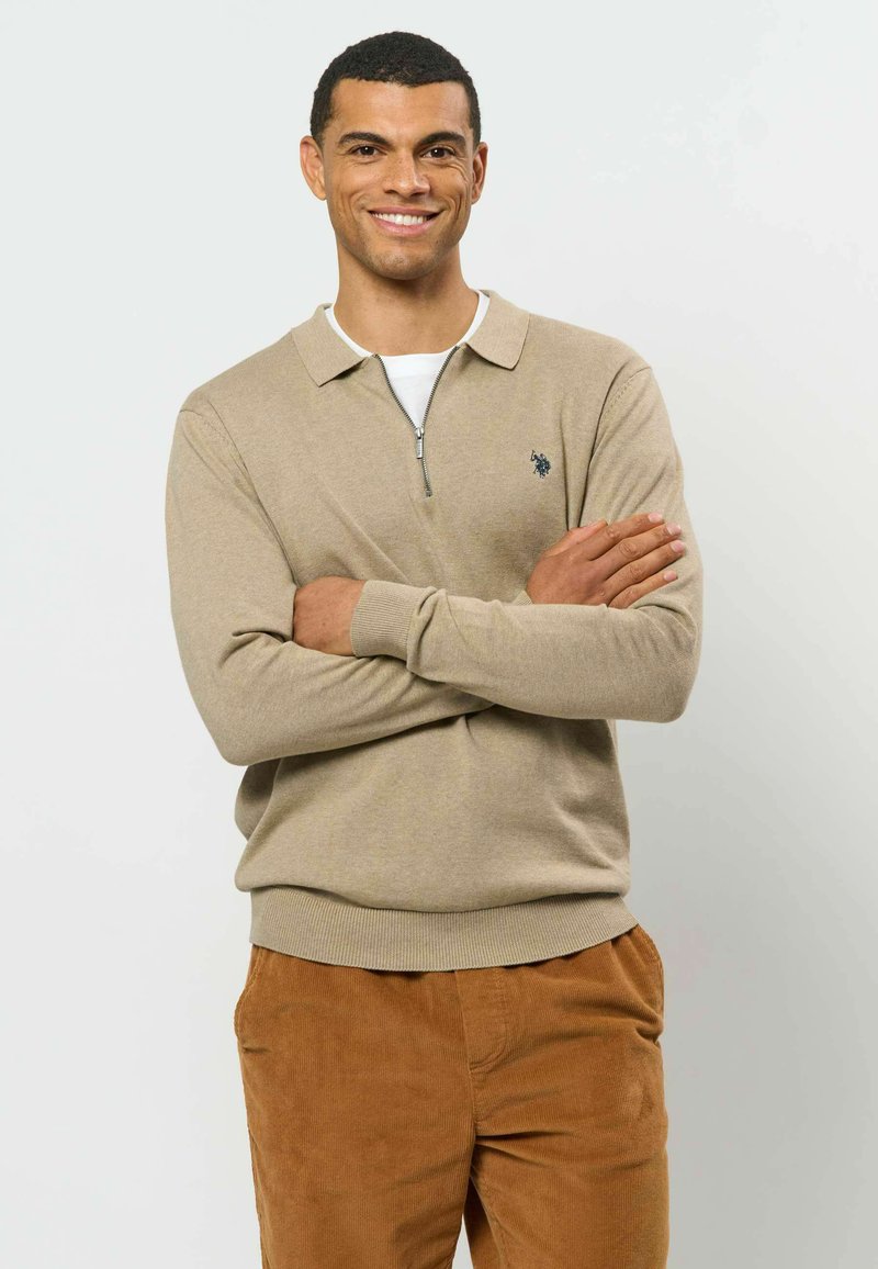 Homme aux cheveux courts et foncés, souriant, les bras croisés, portant un pull beige à col zippé et un pantalon en velours côtelé marron sur un fond uni.