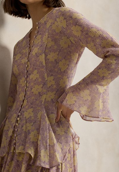 Frau trägt ein langärmliges Kleid mit lila und gelbem Blumenmuster, bekleidet mit stoffbezogenen Knöpfen und ausgestellten Manschetten, posiert mit einer Hand in der Hüfte.