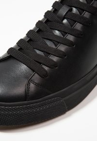 Gros plan d'une basket en cuir noir avec une semelle en caoutchouc texturée noire et des lacets en tissu noir bien serrés sur fond blanc.