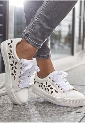 White sneakers with cut-out floral design, flat sole, and white laces. Textured surface and gold eyelets accent the overall look.
