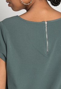 Top verde cazador con tejido texturizado, con mangas cortas y detalle de cremallera en la espalda. El escote es amplio y redondeado.