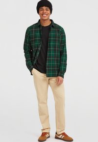 Grønternet flannel skjorte lagd over en sort t-shirt, beige bukser og brune sneakers. Skjorten har en krave med knapper og en brystlomme.