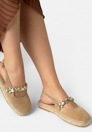 Beige suède slingback espadrilles met edelsteenversieringen op de bandjes, gedragen met een bruine rok op lichtgekleurde benen tegen een witte achtergrond.