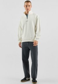 Biała bluza z zamkiem na ćwiartkę i ściągaczami na mankietach, zestawiona z luźnymi, ciemnoszarymi dżinsami oraz czarnymi niskimi trampkami z białymi akcentami.