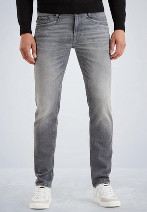 Mann trägt slim fit ausgewaschene graue Jeans, schwarzes Langarmshirt und weiße Sneaker, steht auf grauem Boden.