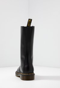 Black leather mid-calf boot with yellow stitching and a black-and-yellow pull tab on a white surface against a plain gray wall.