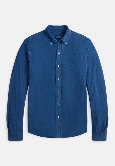 Ilgas rankovėmis mygtukais užsegamas marškinėliai tamsiai mėlynos spalvos audinyje, kuriame matyti subtili faktūra, su mygtukais priekyje ir mažu logotipu kairėje krūtinės pusėje.