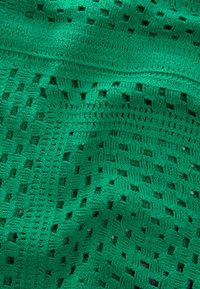 Kootud roheline kangas, mis näitab avatud sooniku mustrit, kus on teineteisega seotud ruudud ja tekstureeritud sooned. Pehme ja painduv, detailse käsitööga.