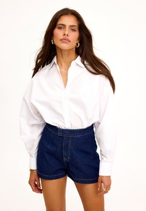 Vrouw die een oversized wit overhemd draagt, ingestopt in een hooggetailleerde donkere spijkershort, met gouden oorringen en ringen.