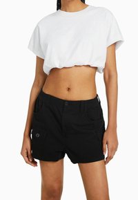 Žena v bílé zkrácené tričku a černých šortkách s vysokým pasem a zapínacími kapsami, stojící proti bílému pozadí.
