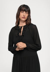 Abito nero con maniche lunghe, scollatura arricciata e chiusura con lacci. Realizzato in tessuto leggero con una gonna fluida.
