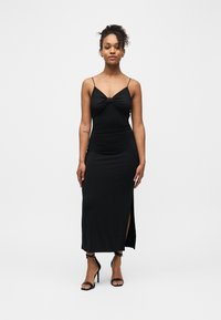 Femme debout portant une robe noire sans manches à fines bretelles, fendue sur le côté, et des sandales à talons hauts noires devant un fond blanc uni.