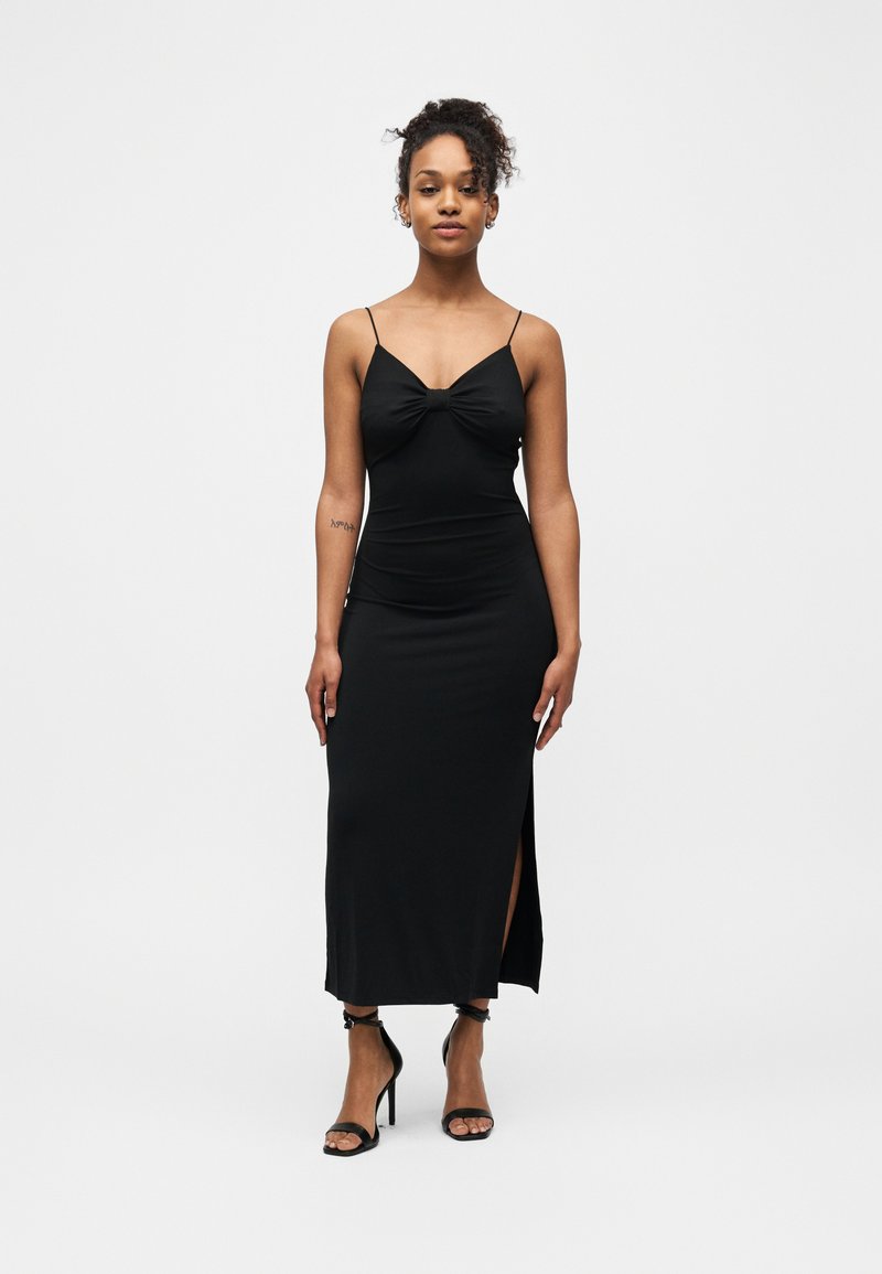 Femme debout portant une robe noire sans manches à fines bretelles, fendue sur le côté, et des sandales à talons hauts noires devant un fond blanc uni.