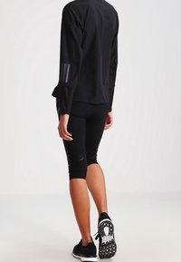 Persoon die met de rug naar de camera staat, gekleed in een zwart sportjack, zwarte caprilengte leggings en zwarte hardloopschoenen met een patroon op de zolen.