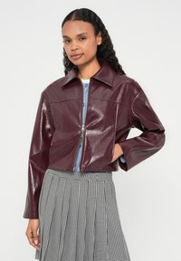 Bordowa kurtka cropped z ekoskóry z zaostrzonym kołnierzem, zapięciem na zamek z przodu i teksturowanym wykończeniem, noszona na niebieskiej bluzce i spódnicy w kratę.