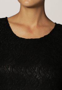 Top de encaje negro con escote redondo, patrón de espiral texturizado y forma ajustada, que exhibe una mezcla de elegancia y simplicidad.