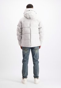 Grijze pufferjack met een capuchon, met brede horizontale stiksels en een logo op de achterkant. Gecombineerd met vervaagde blauwe jeans en witte sneakers.