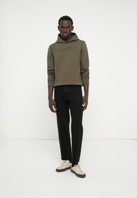 Haut à capuche vert olive avec une poche kangourou et impression de logo, associé à un jean droit noir et des baskets crème avec des semelles marron.