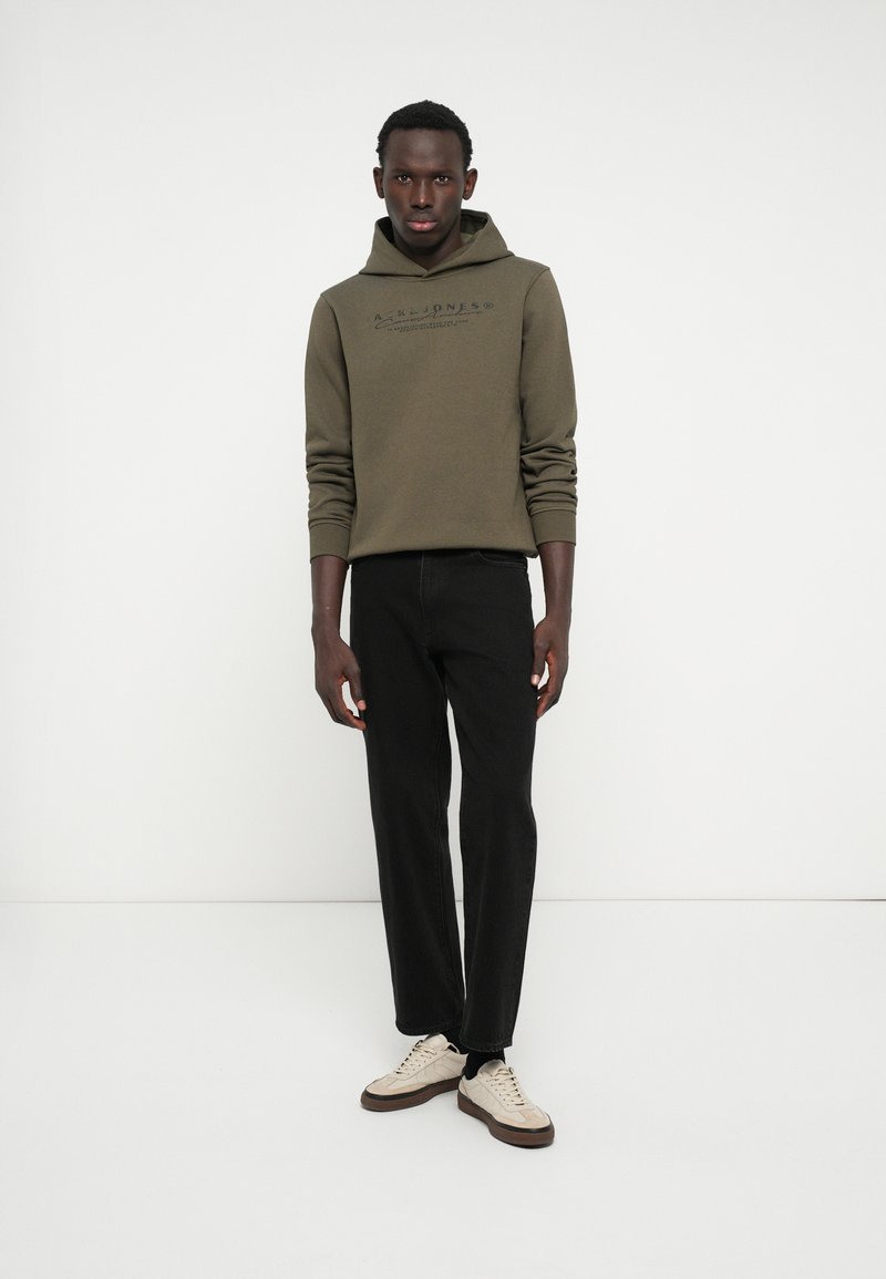 Haut à capuche vert olive avec une poche kangourou et impression de logo, associé à un jean droit noir et des baskets crème avec des semelles marron.