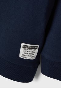 Sudadera azul marino con puños acanalados texturizados. Una etiqueta blanca muestra el texto con las instrucciones de cuidado y detalles del producto.