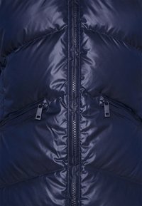 Marinblå pufferjacka med en slät, blank yta, utrustad med en central dragkedja, två dragkedjefickor och subtila diagonala sömmar.