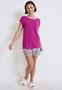 Top de manga corta en fucsia combinado con pantalones cortos estampados con diseños florales en verde y rosa. La modelo lleva pantuflas blancas y esponjosas.