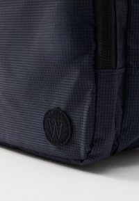 Navy blue fabric backpack with a grid pattern. Features a textured, circular black patch with a logo, and durable zip closures.