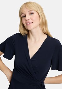 Robe portefeuille bleu marine en tissu doux, avec des manches courtes et flottantes et un lien à la taille. Texture lisse et décolleté en V.