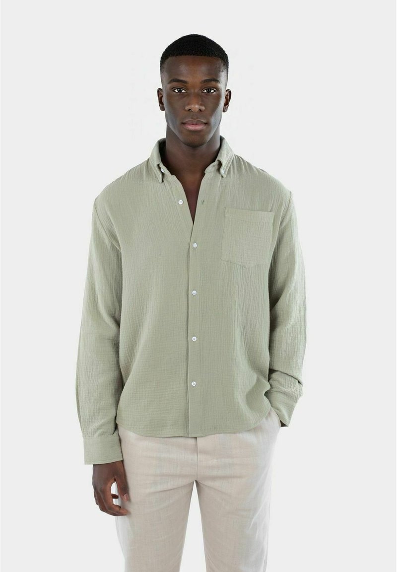 Camicia button-up verde chiaro con un tessuto testurizzato, taschino sul petto e maniche lunghe. Abbinata a pantaloni di colore chiaro.