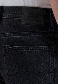 Close-up van een zwarte denim jeanszak met een zwarte Lee-merkpatch en een klein geel Lee-label aan de zakrand.