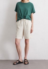 Camiseta verde de manga corta, de corte holgado, combinada con pantalones cortos de lino crema con cintura elástica. Sandalias negras con detalles de hebilla dorada.