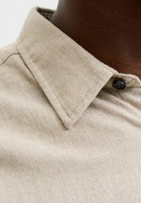 Beige collared shirt buttoned at the neck on dark brown skin, showing detailed fabric texture and stitching.