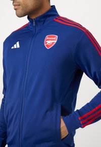 Mėlyna sportinė striukė su raudonais akcentais, turinti užtrauktuką, aukštą apykaklę ir Adidas logotipą dešinėje krūtinės pusėje. Arsenal emblema kairėje.