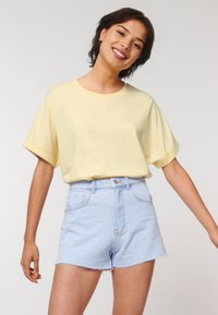 Smiling person with short dark hair, wearing a light yellow t-shirt and light blue denim shorts, poses against a plain background.