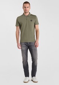 Olivengroene polo shirt met een kraag, drie knopen en een ronde zwart logo. Gecombineerd met grijze jeans en witte sneakers.