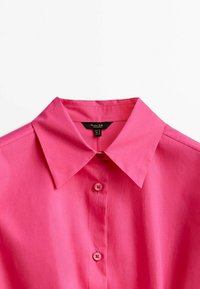 Lys pink skjorte med krave og en glat tekstur, med en knappet front og to synlige knapper. Etiketten siger "Mango."