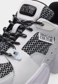 Λευκό και μαύρο αθλητικό παπούτσι Versace Jeans Couture με υφασμάτινα πάνελ με υφή, επώνυμα ετικέτες και κορδόνια σε αντίθετους μαύρους και λευκούς χρωματισμούς.