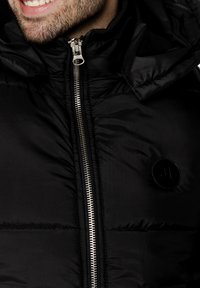 Czarna kurtka puchowa z zamkiem błyskawicznym z przodu i dużym kapturem. Gładki materiał o błyszczącym wykończeniu i okrągłym logo po lewej stronie.