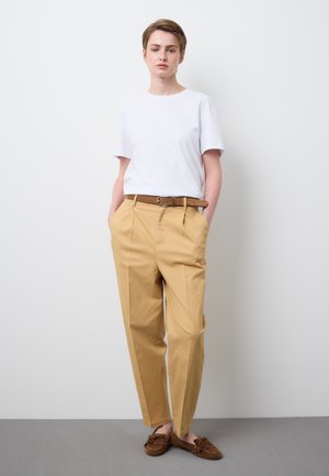 Persona con cabello corto que lleva una camiseta blanca, pantalones beige de talle alto con un cinturón marrón y mocasines marrones, de pie contra una pared blanca.