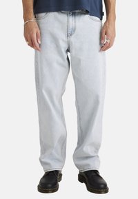 Lichtblauwe denim jeans met een relaxed fit, voorzien van contrasterende stiksels, grote zakken en opgerolde manchetten. Draagt met zwarte schoenen.
