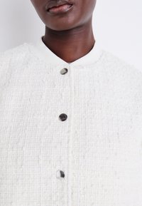 White textured jacket with a woven pattern, featuring a solid white collar and three metallic buttons along the front.