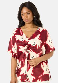 Blusa floreale rossa e crema con scollo a V, maniche corte, dettaglio arricciato sul davanti e accenti neri su un tessuto leggero.