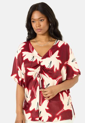 Rote und cremefarbene Bluse mit floralem Muster, V-Ausschnitt, kurzen Ärmeln, gerafftem Vorderteil und schwarzen Akzenten auf leichtem Material.