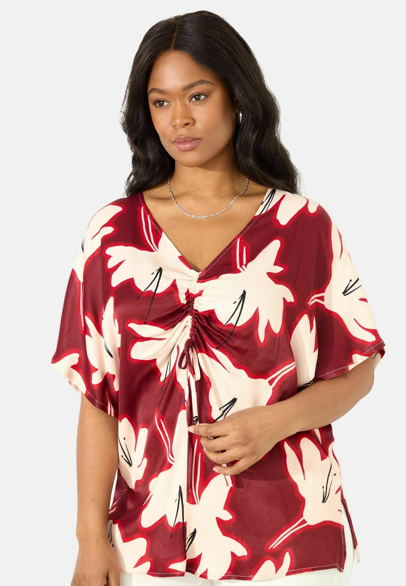 Blusa floreale rossa e crema con scollo a V, maniche corte, dettaglio arricciato sul davanti e accenti neri su un tessuto leggero.