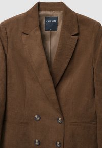 Blazer in corduroy marrone con un davanti doppiopetto, rever a punta e due grandi bottoni. Include una tasca singola sul lato sinistro.