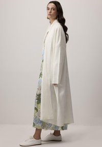 Dlhý, biely kardigan z mäkkej pleteniny, s širokými rukávmi a voľným strihom, nosený cez kvetinové šaty a biele topánky.